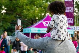 Man with child on shoulders captures selfie in lively outdoor festival setting.